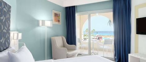 A hotel room with a bed, a chair, a desk, and a TV. There is a window with a view of the beach and palm trees.