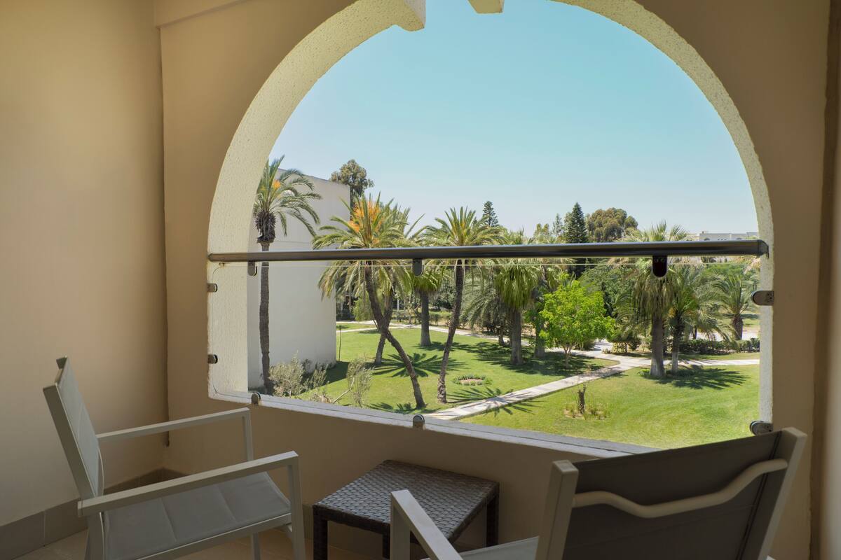 A balcony with a view of a well-maintained garden, a small table, and two chairs.