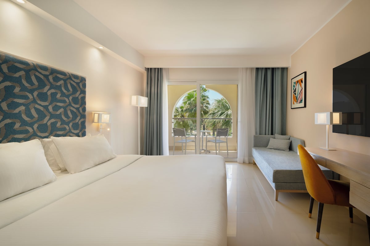 A hotel room with a bed, a desk, a chair, a TV, and a balcony with a view of palm trees.