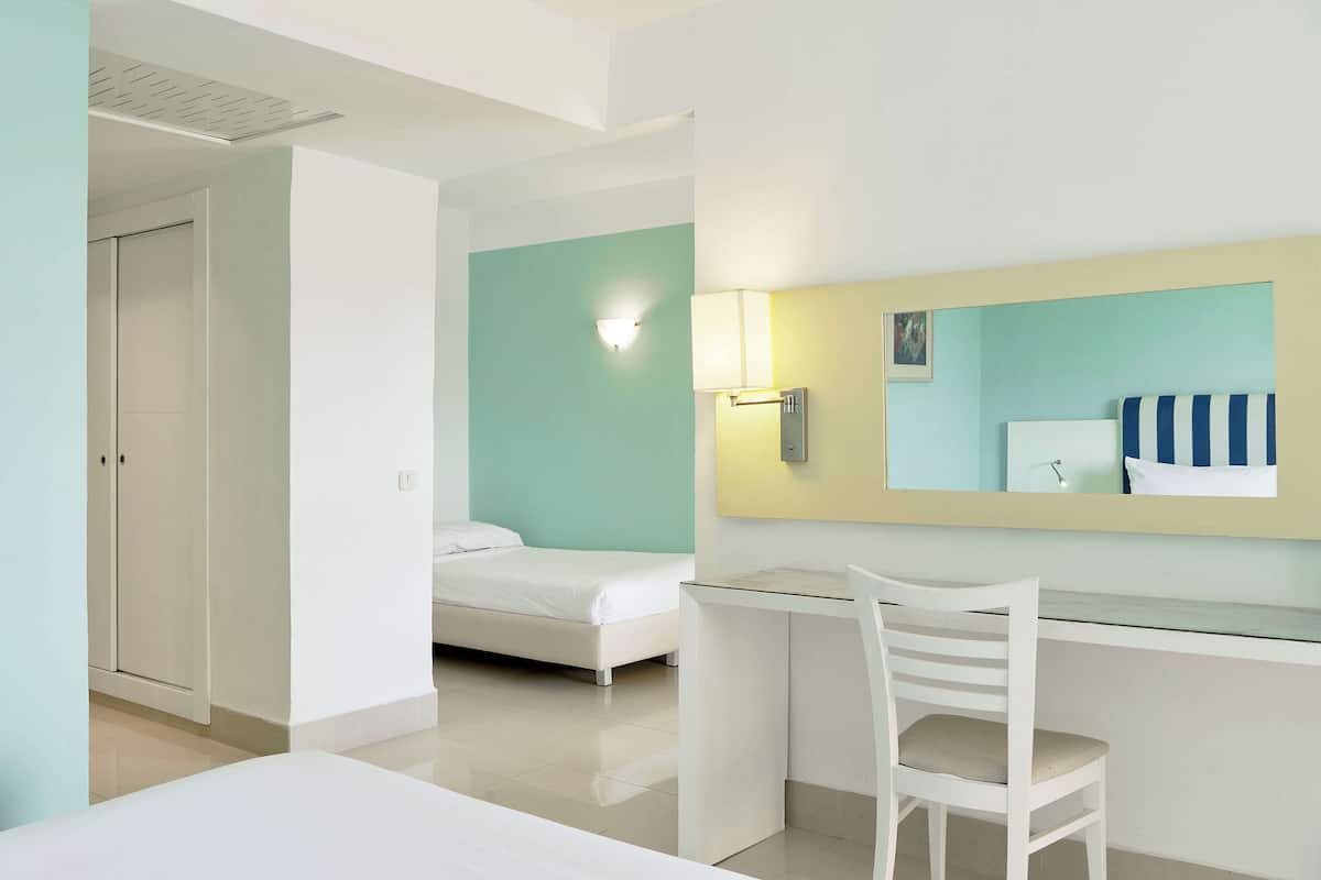 A hotel room with a bed, a desk with a chair, a mirror, and a wall-mounted lamp.