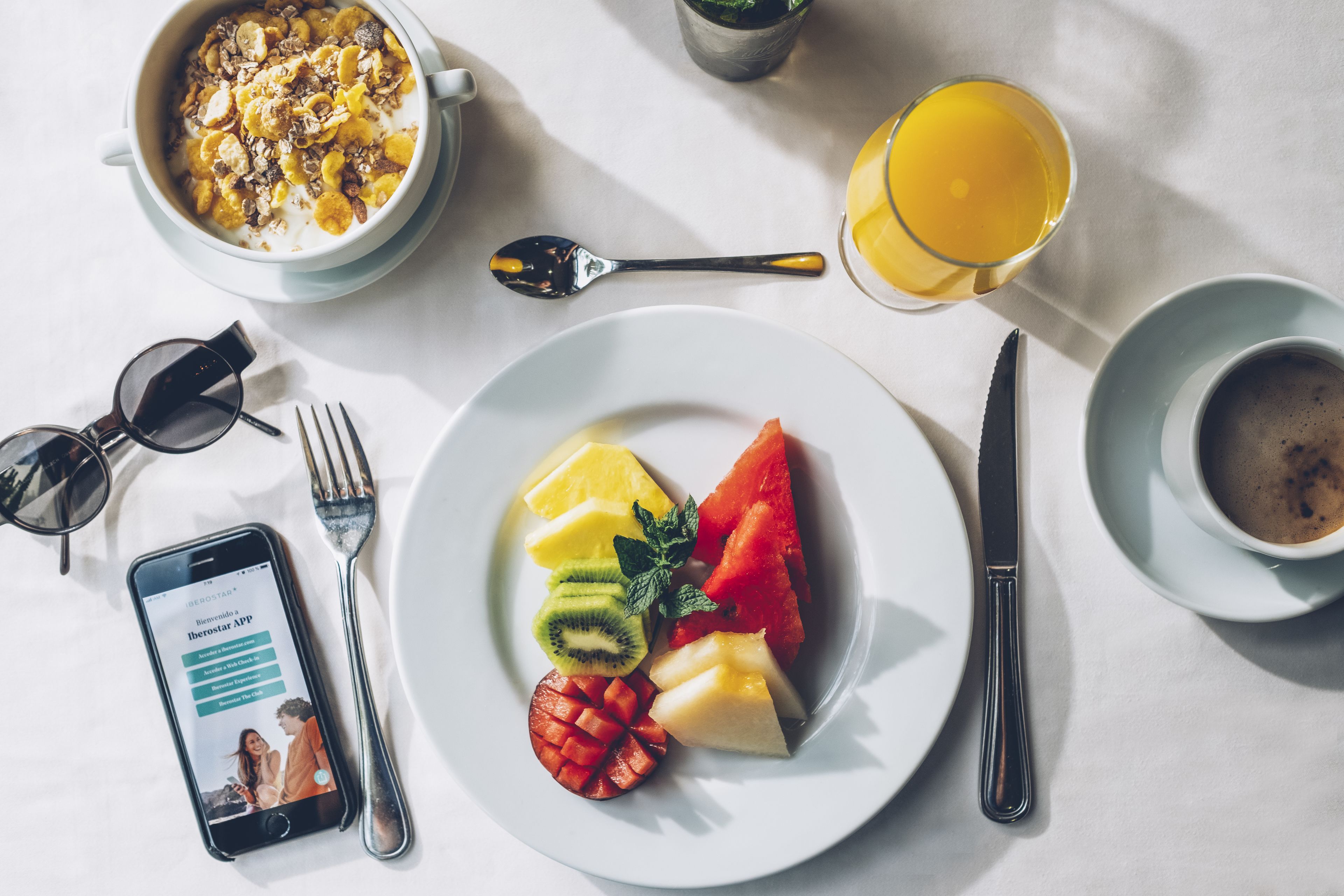 A breakfast setting with a plate of fruit, a bowl of cereal, a glass of orange juice, and a cup of coffee.