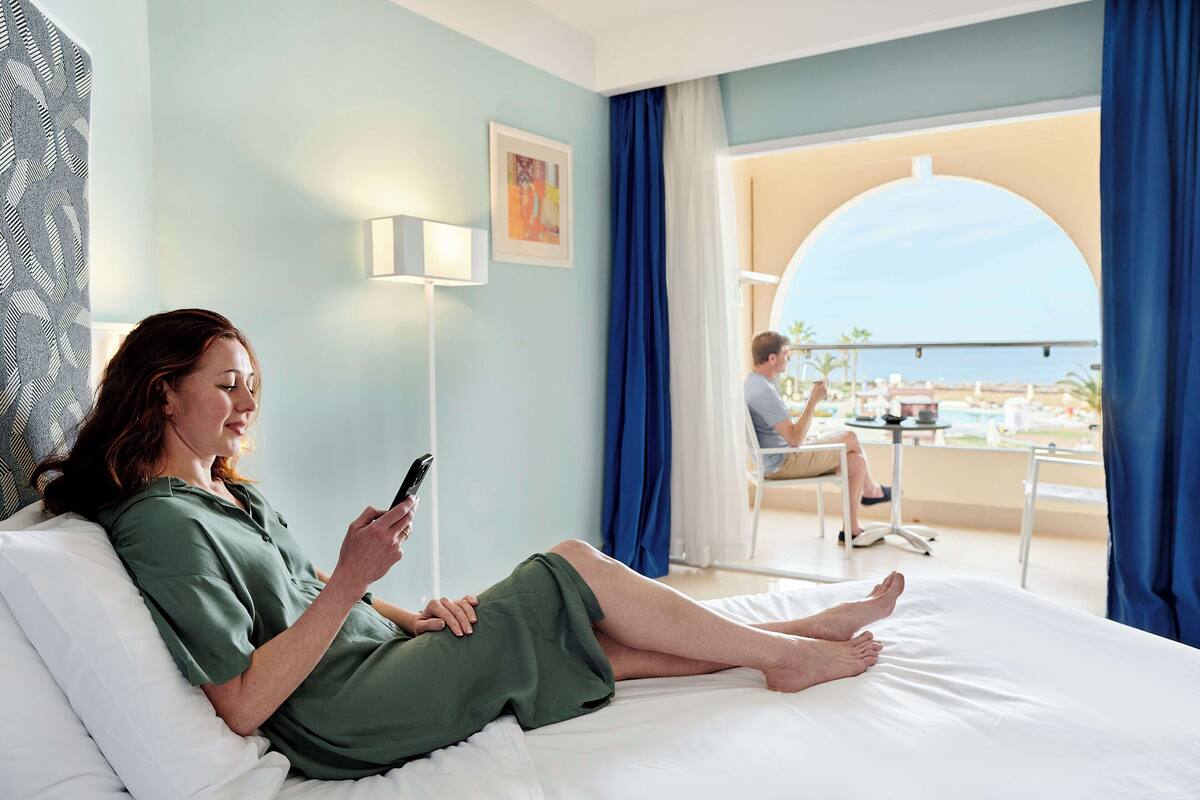A hotel room with a bed, a bedside lamp, a window with curtains, and a view of a pool and beach.