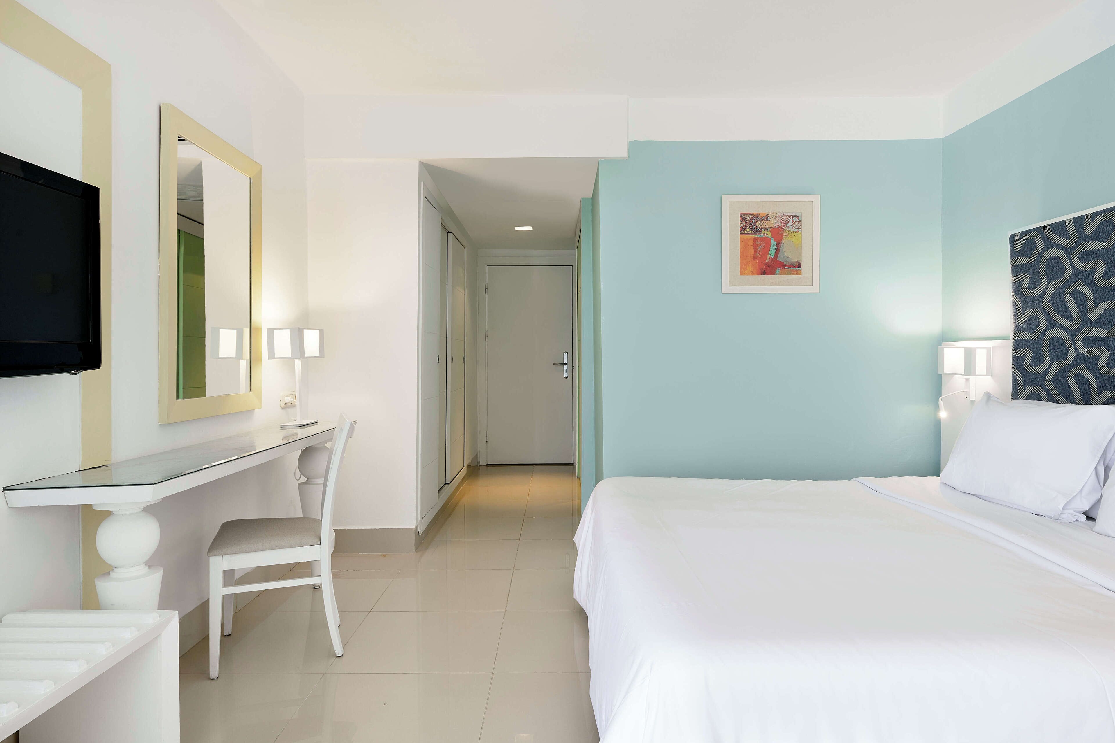 A hotel room with a bed, a desk with a chair, a television, and a door leading to another room.