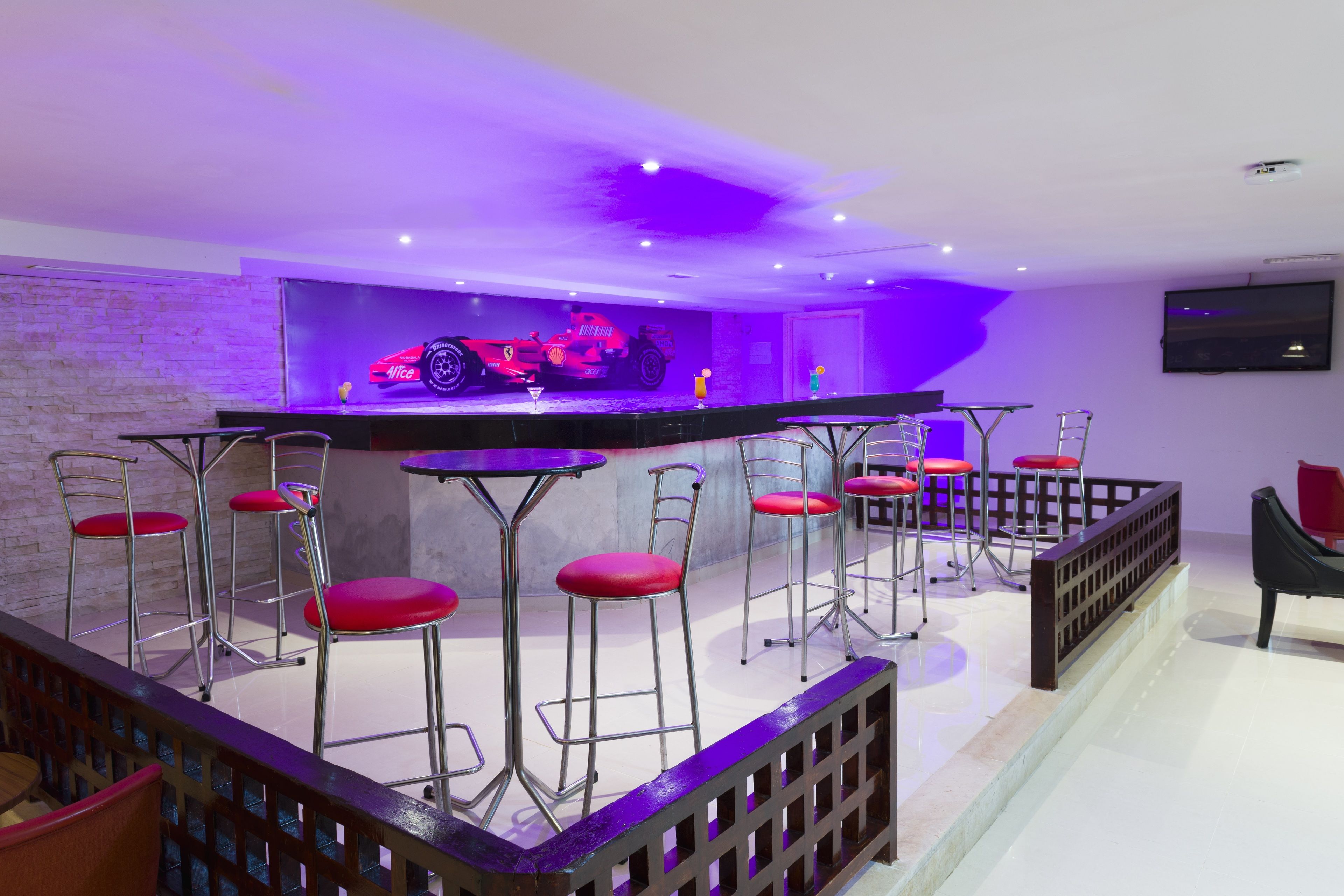 A bar area with red bar stools, a large screen displaying a car, and a television on the wall.