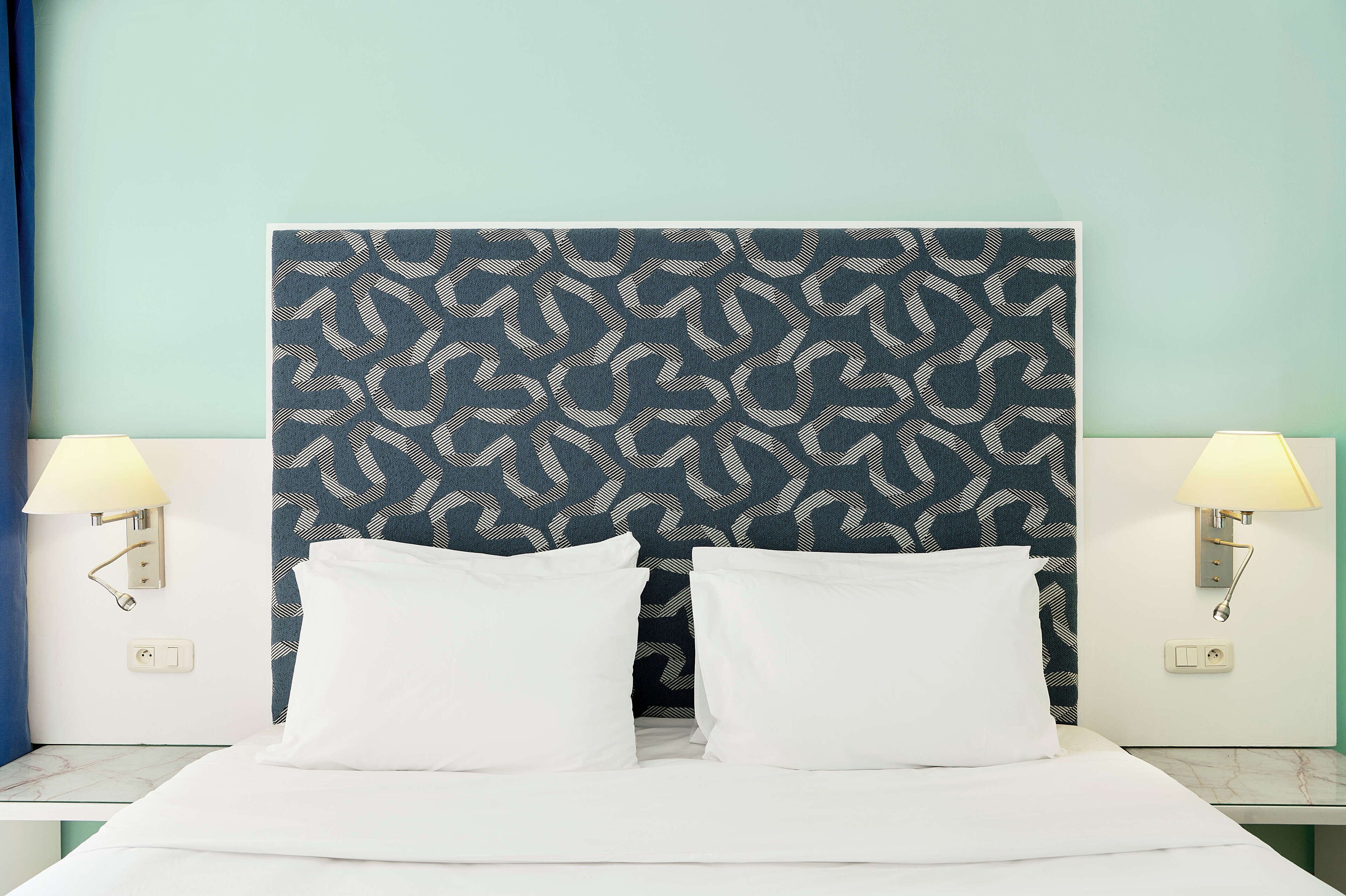 A hotel room with a patterned headboard, two white pillows, a bed, and two wall-mounted lamps.