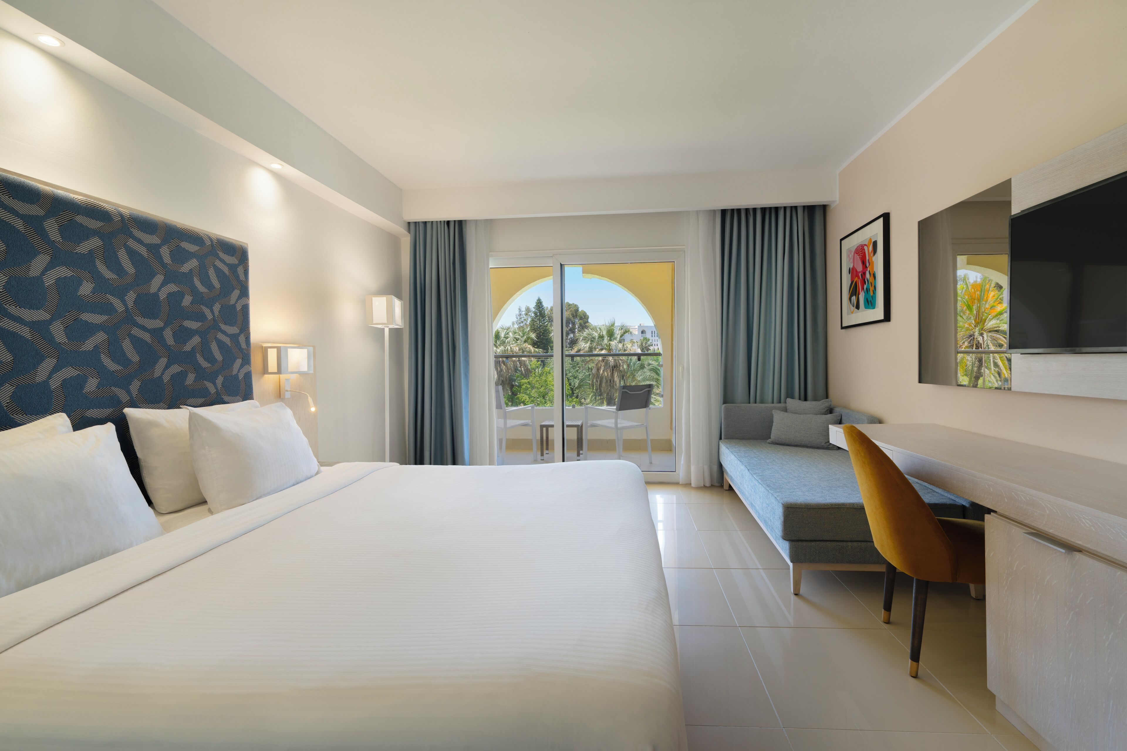A hotel room with a large bed, a desk, a chair, a sofa, and a balcony with a view.