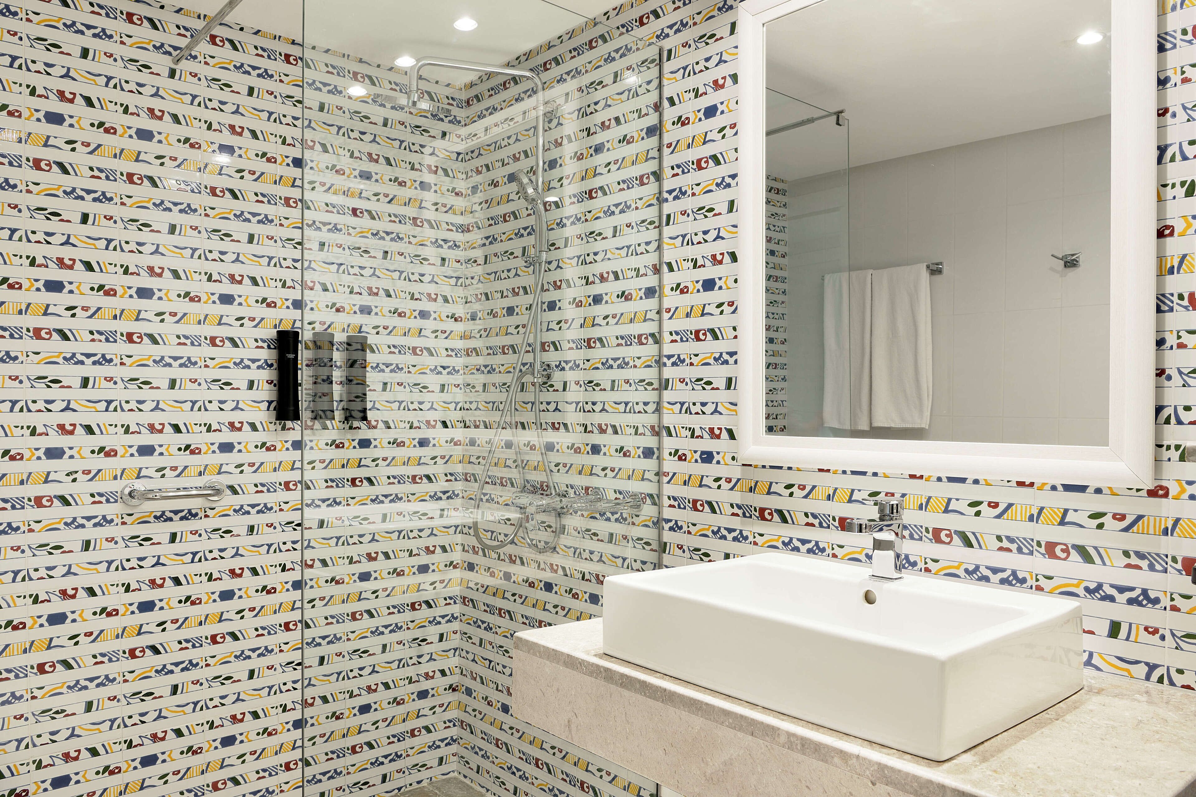 A bathroom with a tiled shower wall, a large mirror, and a white sink.