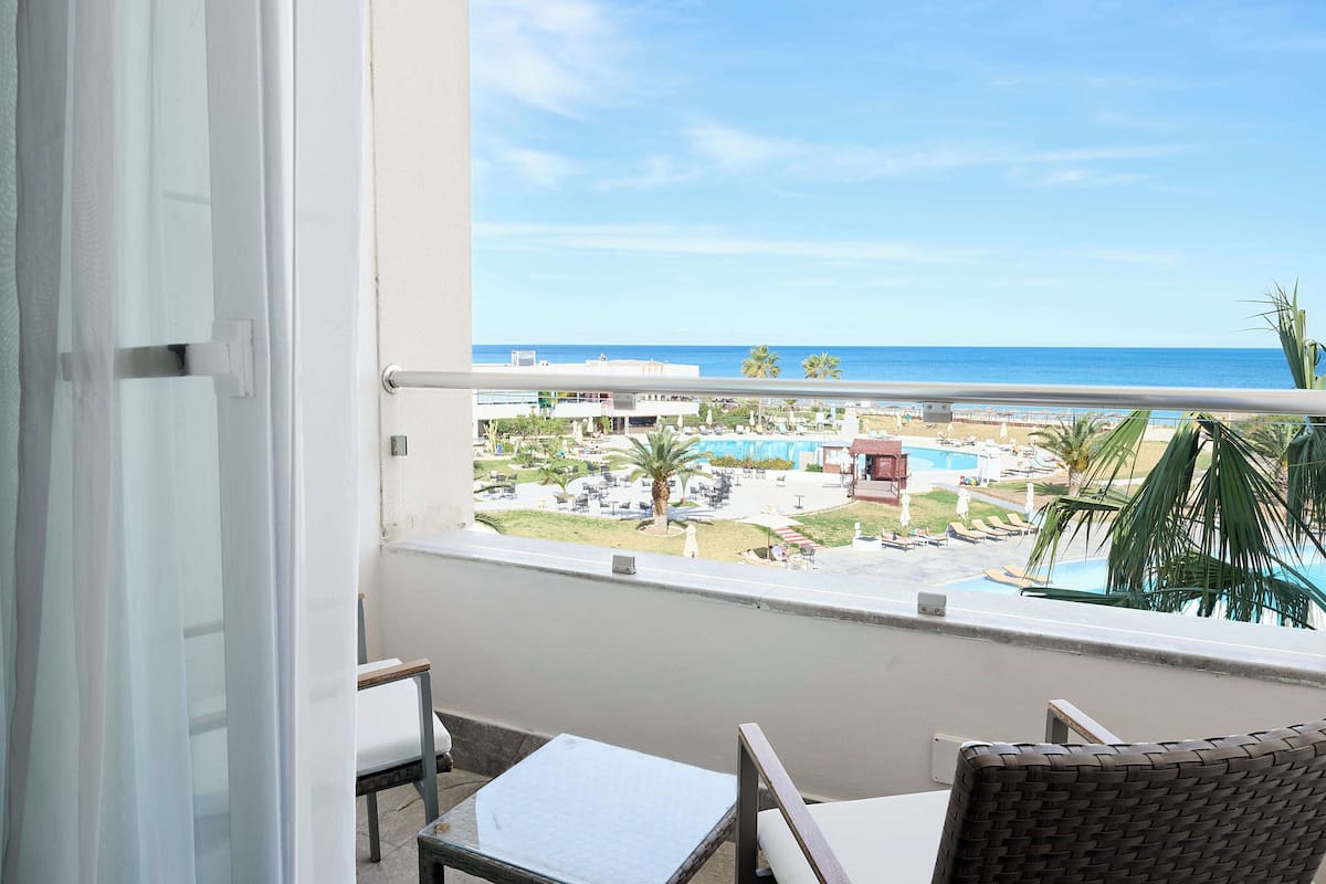 A balcony with a view of a pool and beach, a chair, and a table.