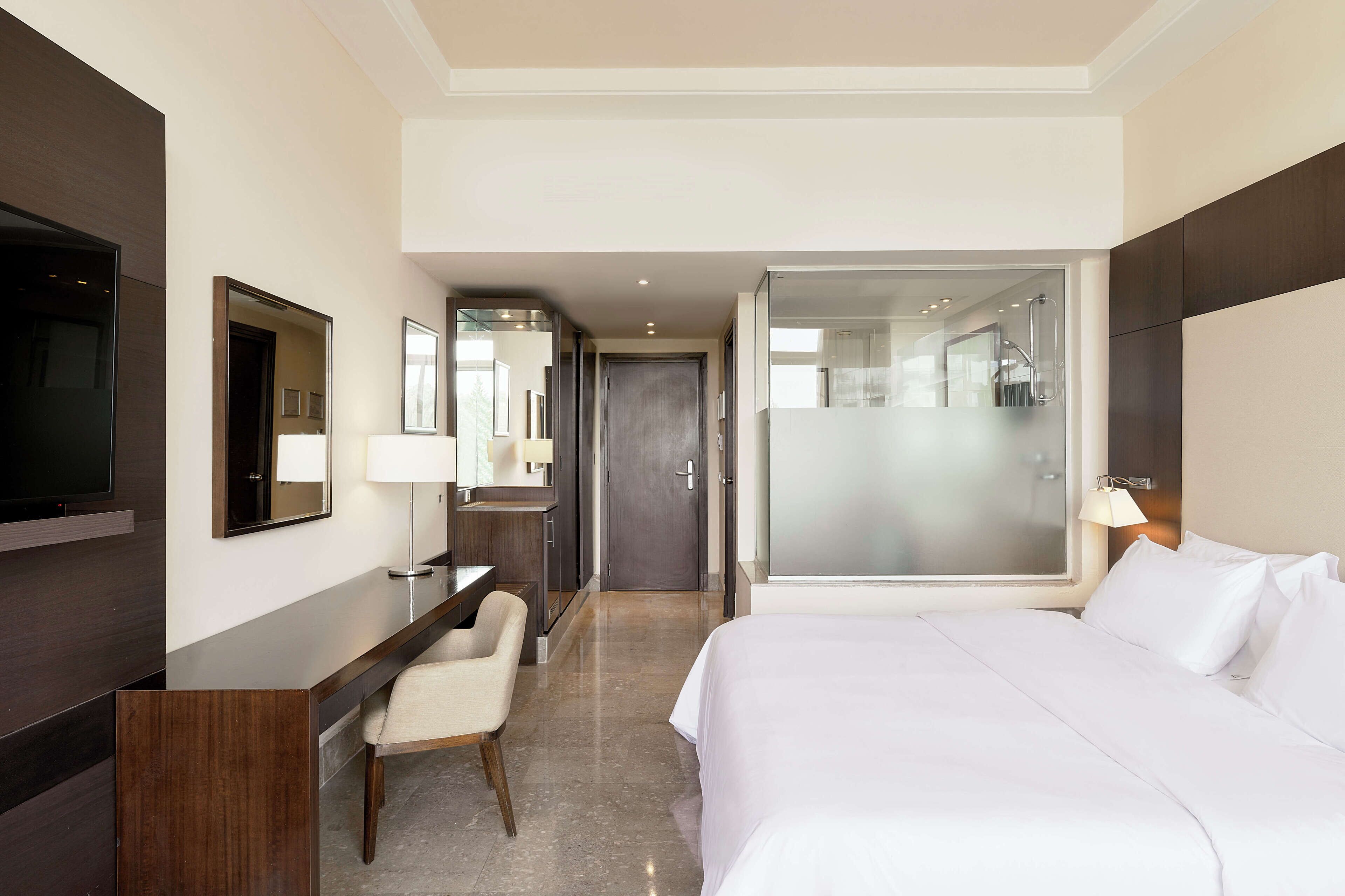 A hotel room with a bed, a desk, a chair, a TV, and a bathroom visible through a glass door.