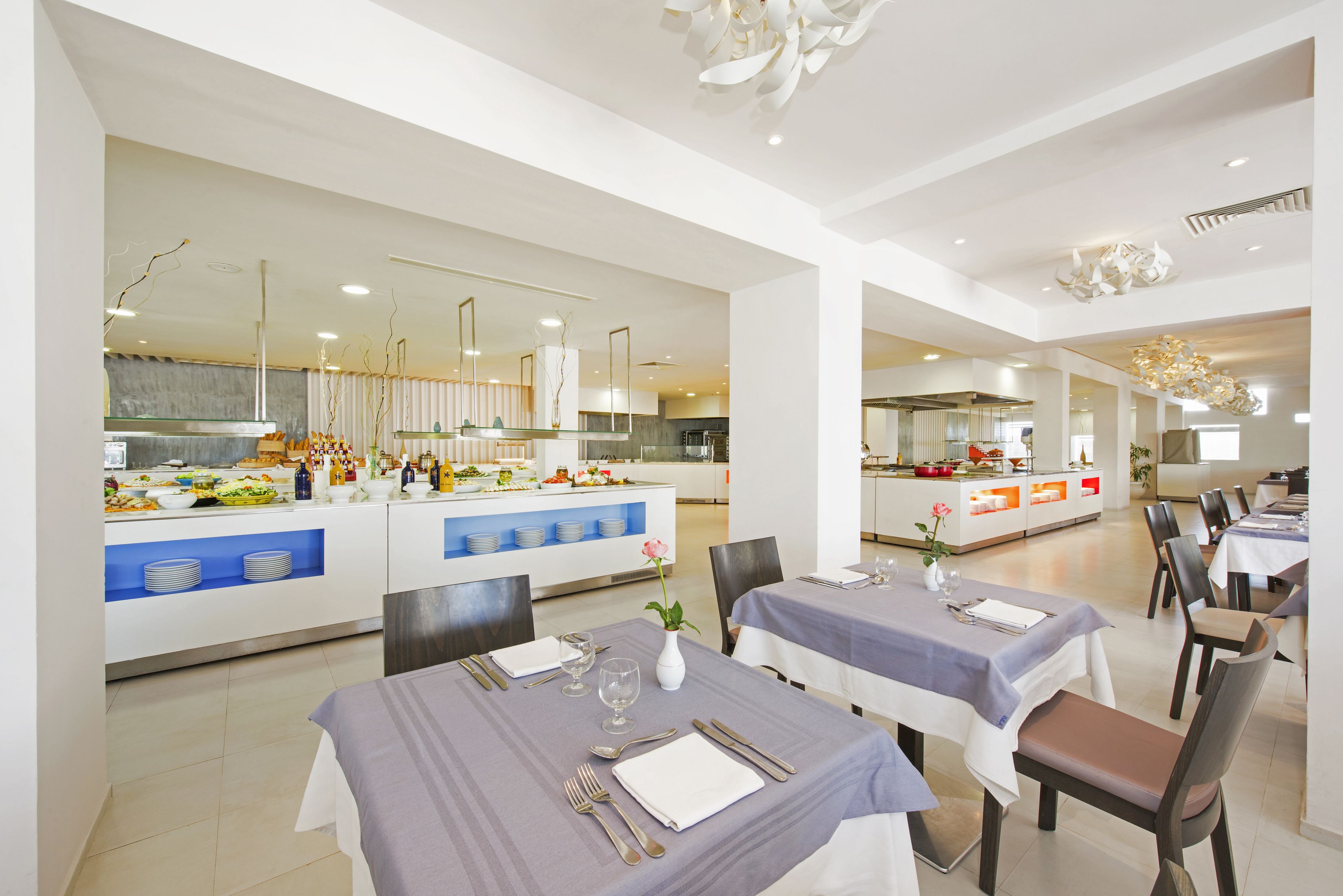 A modern dining area with tables set for a meal, a buffet with various food items, and a kitchen in the background.
