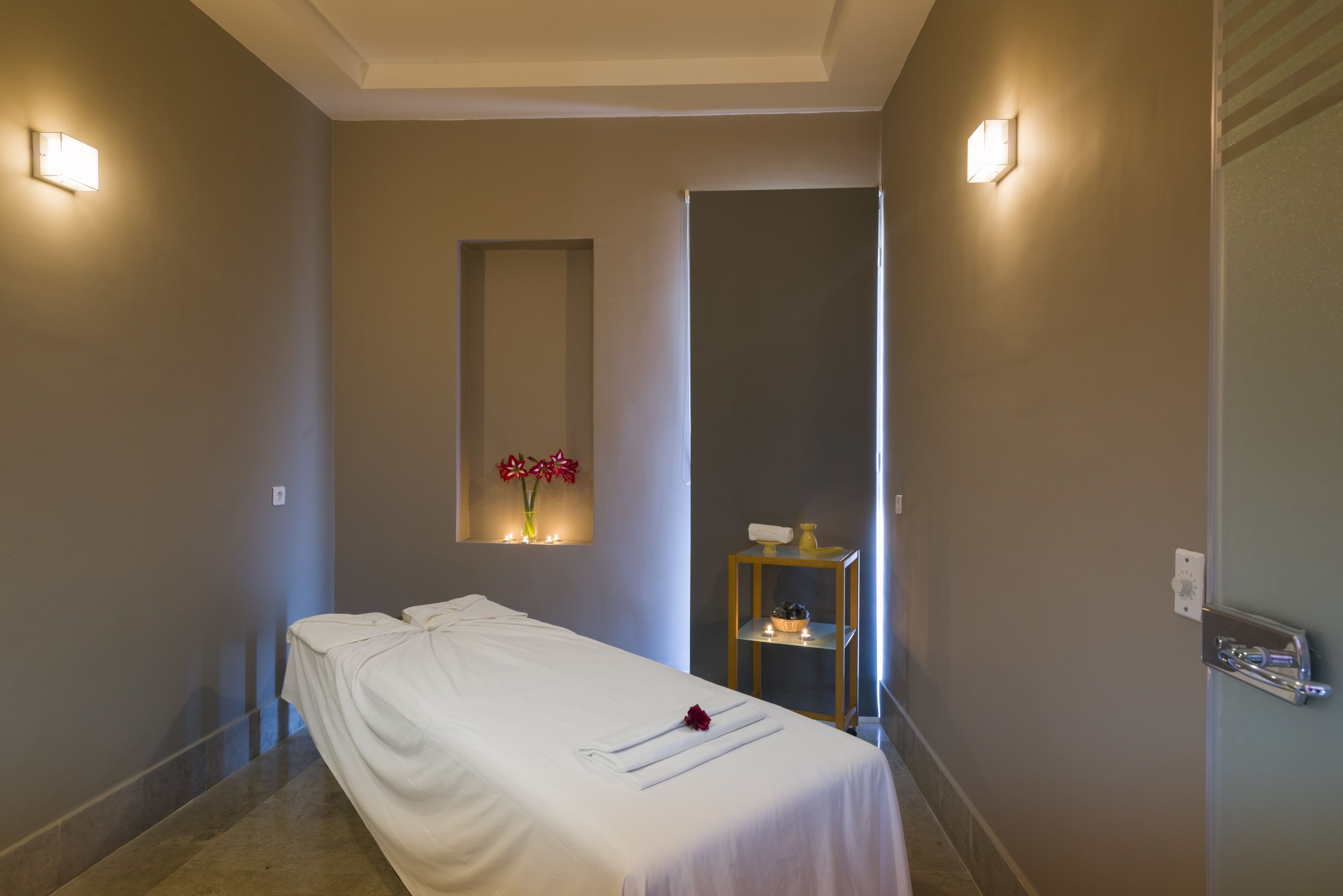 A single bed with a white bedspread, a vase with red flowers, and a small table with a candle.