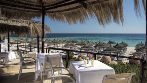Un espace de restauration en plein air, avec des tables et des chaises, donnant sur une plage agrémentée de parasols et de chaises longues.