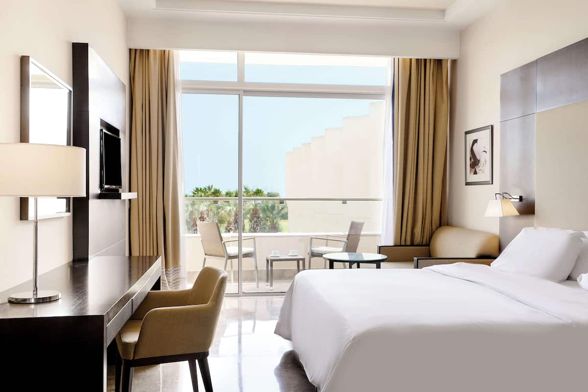 A hotel room with a bed, a desk with a TV, a chair, a small table, and a balcony with curtains.