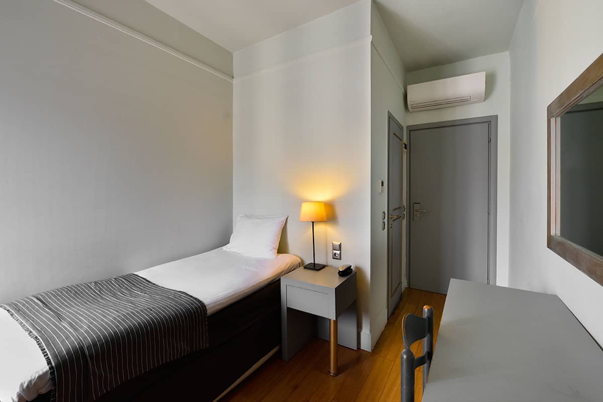 Ein kleines Zimmer mit einem Einzelbett, einem Nachttisch, einer Lampe, einem Stuhl und einem Schreibtisch.