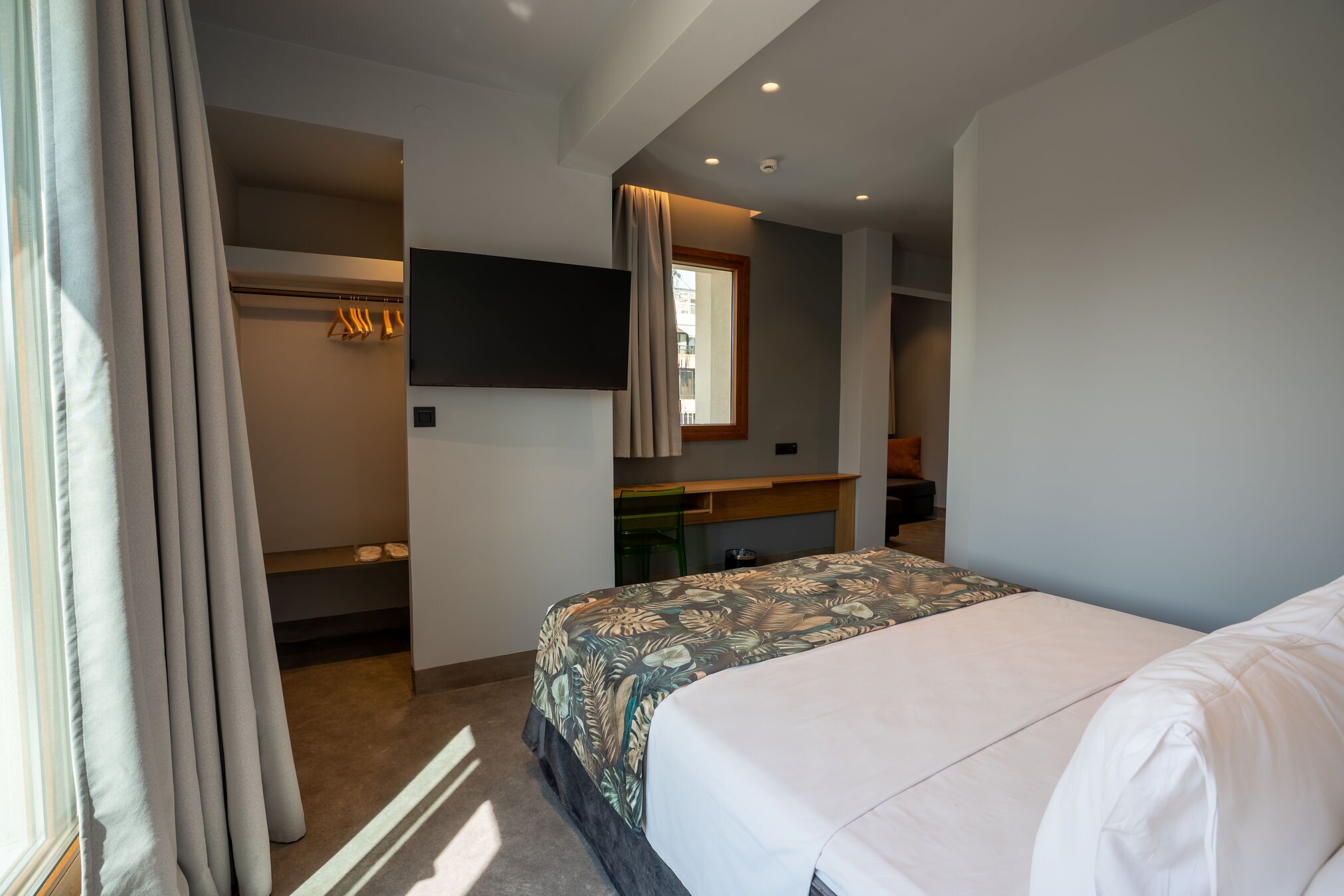 A bedroom with a bed, a desk, a chair, a TV, and a window.