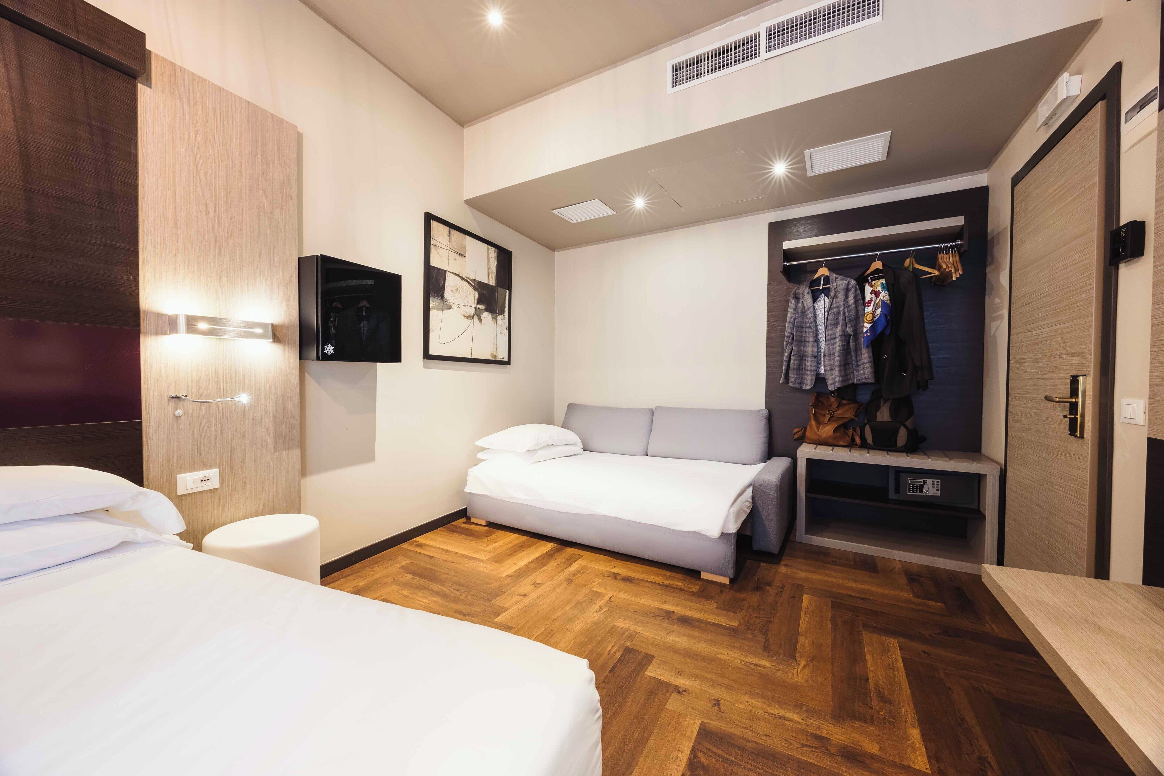 Ein modernes Hotelzimmer mit Holzparkett, einem Bett, einem Nachttisch, einem Kleiderschrank und einer kleinen Sitzecke mit einer Bank.