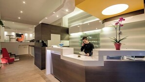 A modern reception area with a receptionist, a reception desk, a potted orchid, and a staircase in the background. - Genius Hotel Downtown (Milan)