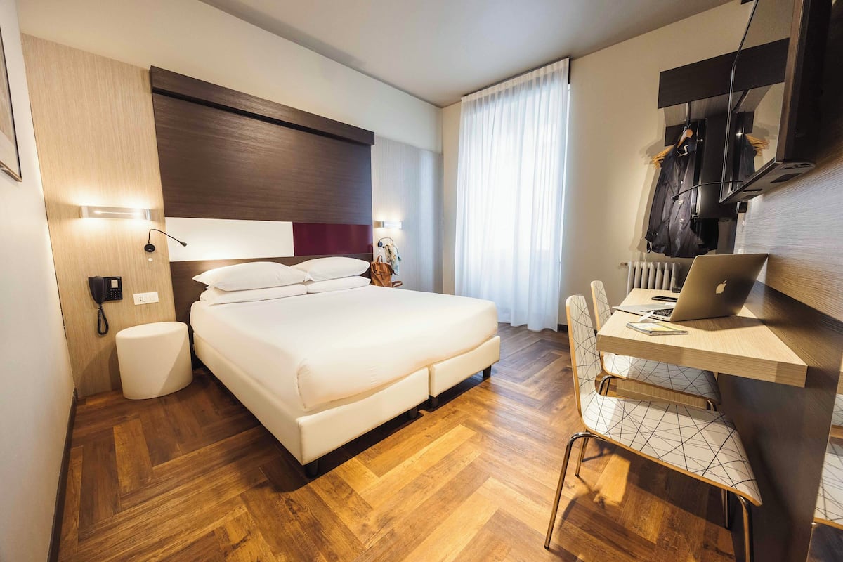 A modern hotel room with a large bed, a desk with a laptop, a chair, and a radiator.