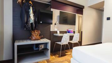 A modern hotel room with a wooden herringbone floor, a bed, a desk with a chair, a TV mounted on the wall, and a coat rack with clothes and a handbag.