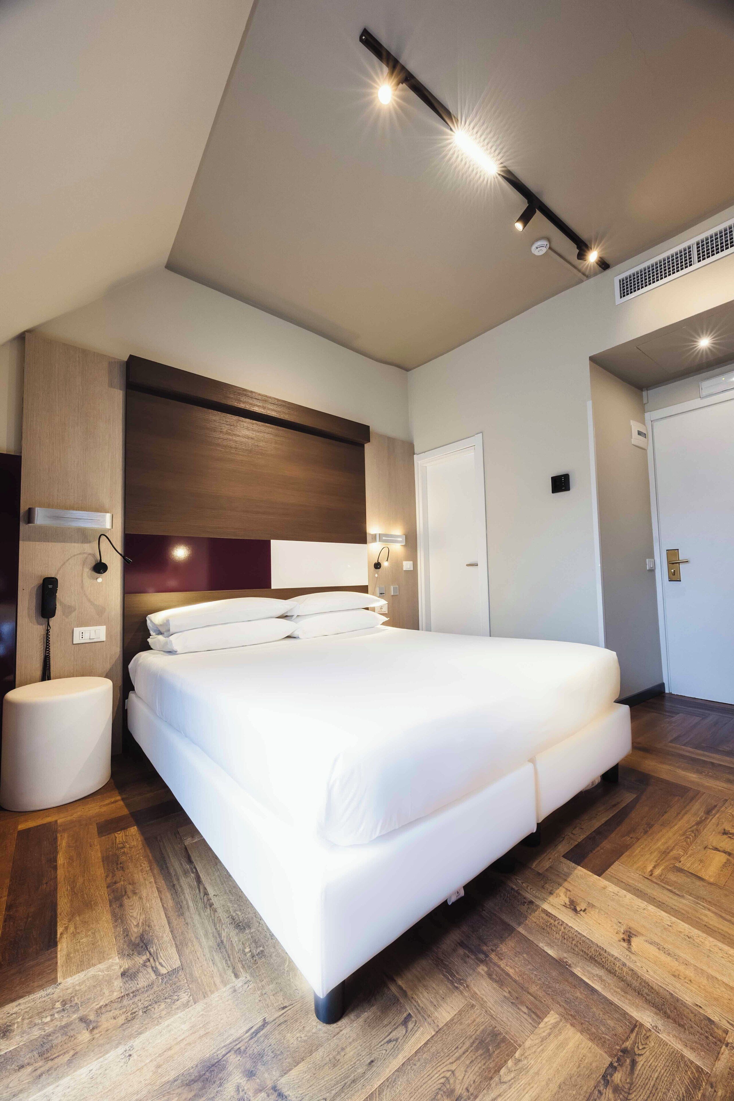 A modern hotel room with a large bed, wooden flooring, and built-in lighting.