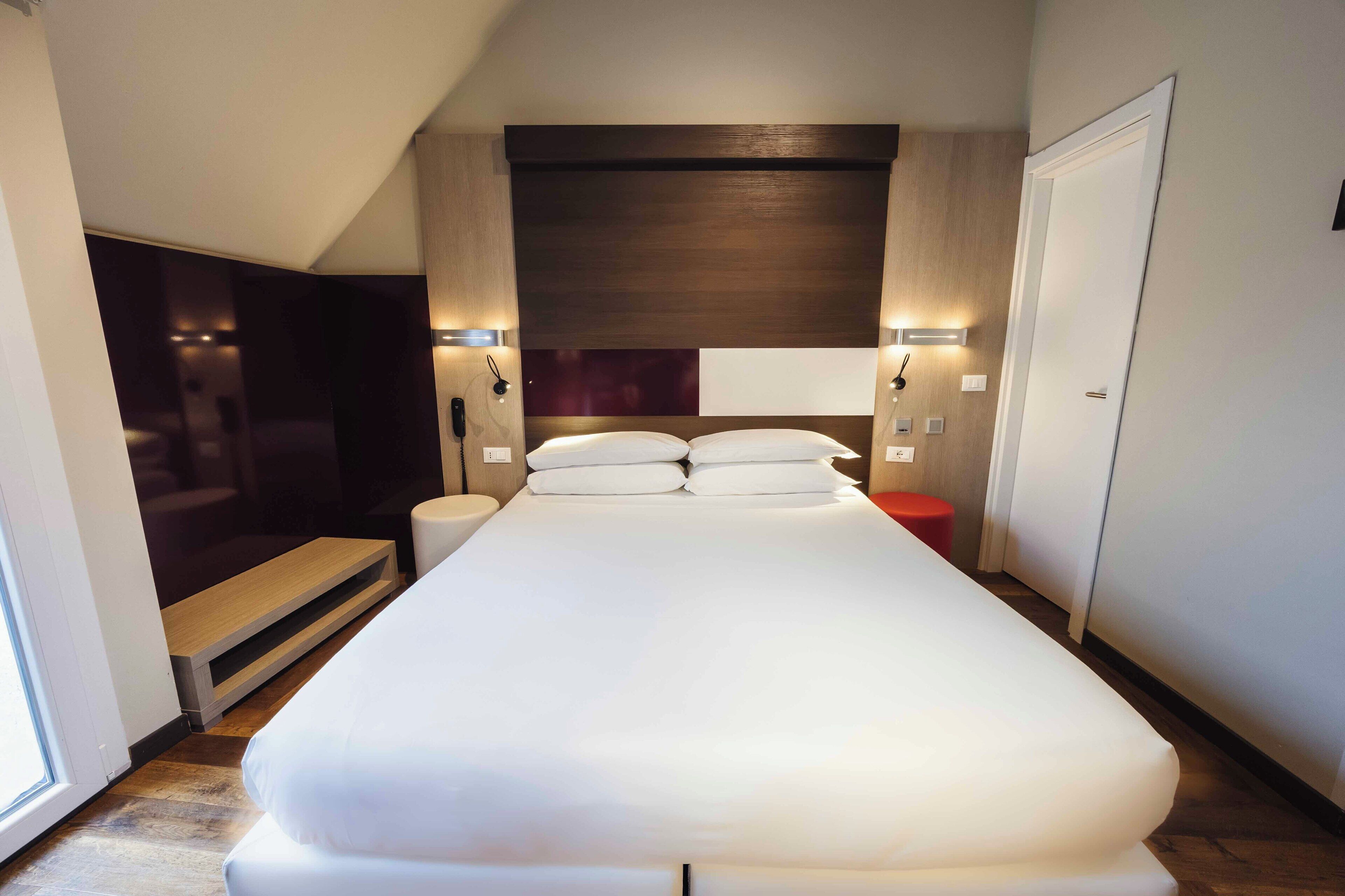 Una habitación de hotel moderna con una cama grande, mesitas de noche, televisión integrada y un taburete rojo.
