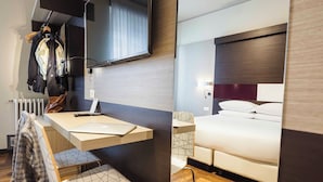A modern hotel room with a wooden floor, a bed with white bedding, a bedside table with a lamp, a chair, a desk with a laptop, and a large mirror reflecting the room. - Genius Hotel Downtown (Milan)