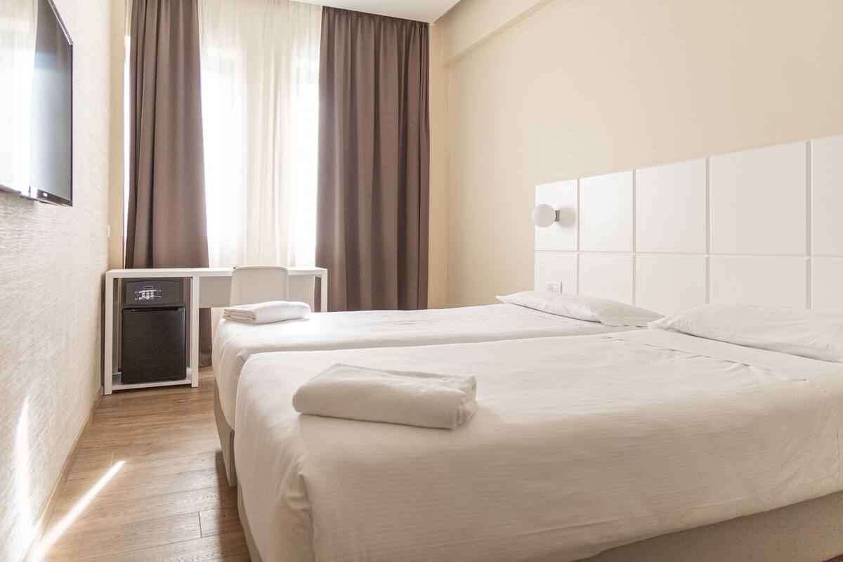 Une chambre d’hôtel avec deux lits, un bureau, un petit réfrigérateur et un téléviseur fixé au mur.