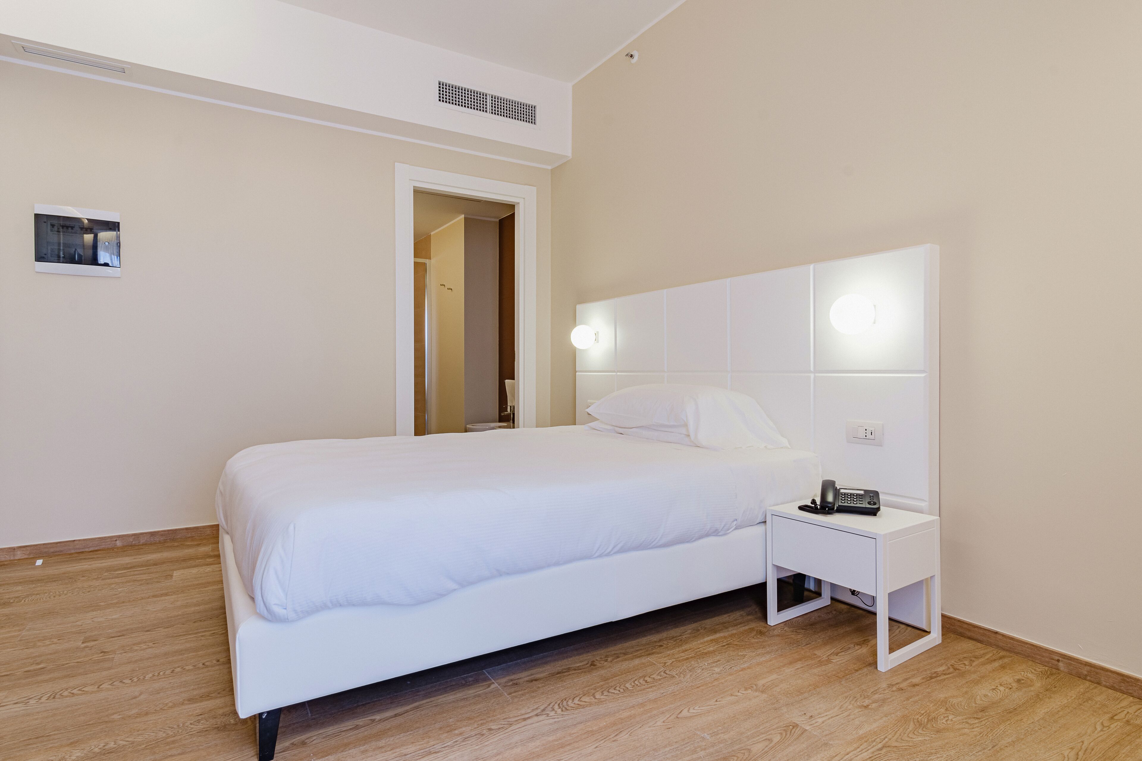 A bedroom with a white bed, a bedside table with a phone, and a door leading to another room.