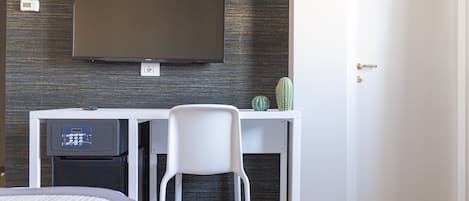 A hotel room with a flat-screen TV mounted on the wall, a white desk with a chair, a small cactus, and a door.