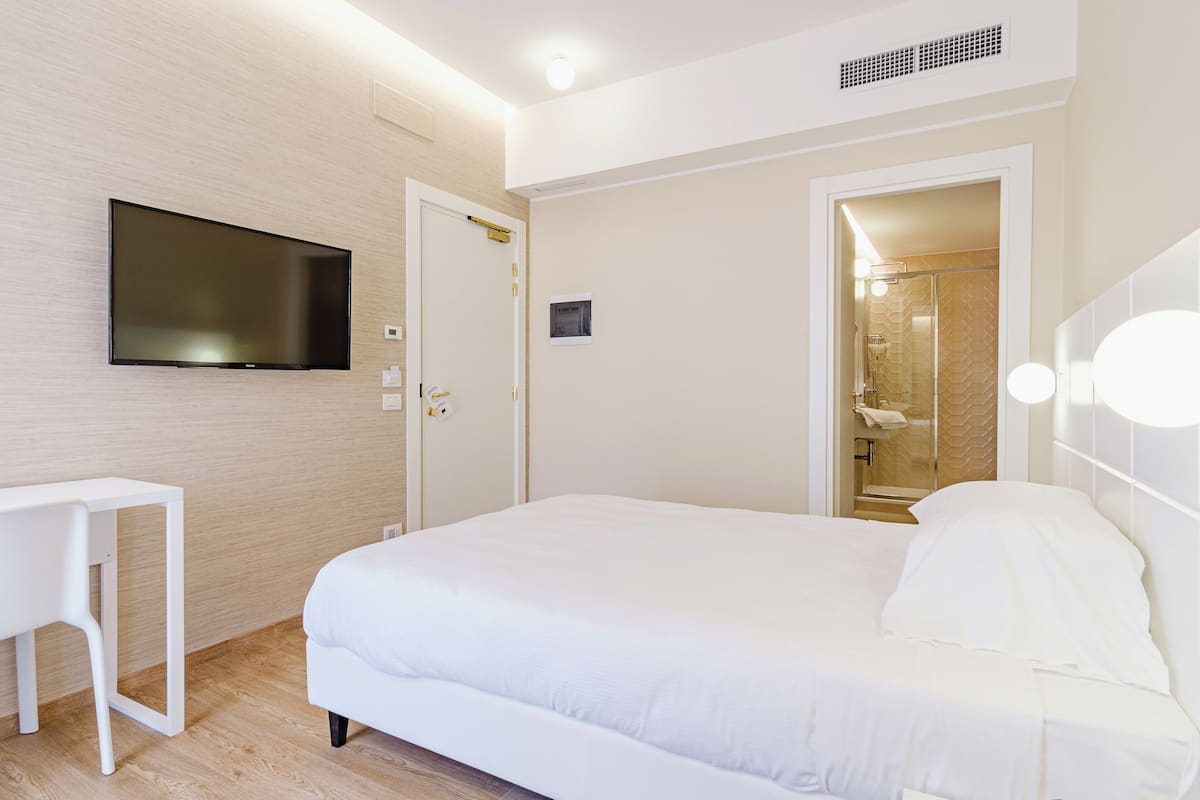 Une chambre d’hôtel moderne dotée d’un grand lit, d’un téléviseur à écran plat fixé au mur, d’une petite table et d’une douche à l’italienne avec une porte en verre.
