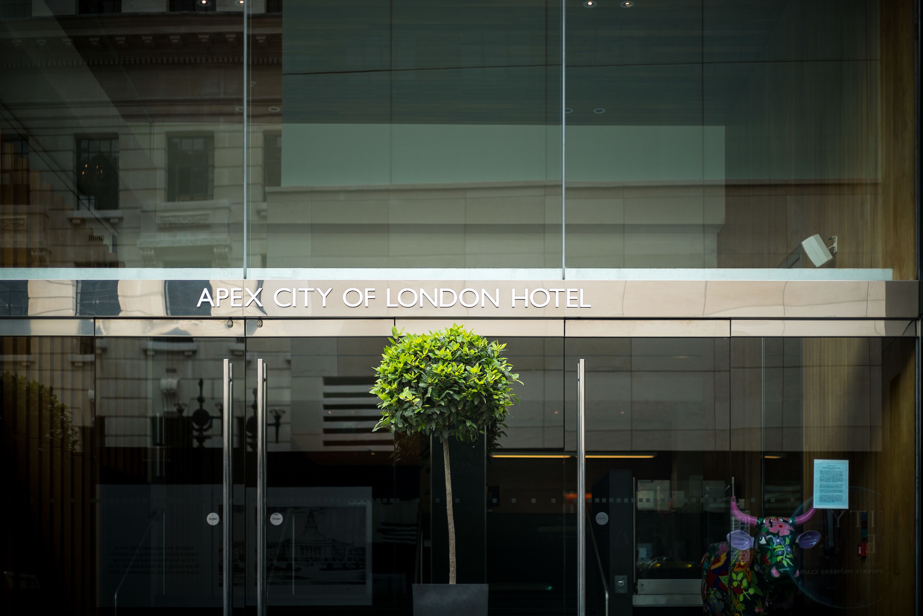 The exterior of the Apex City of London Hotel with a modern glass facade, a potted plant, and a decorative bull sculpture.
