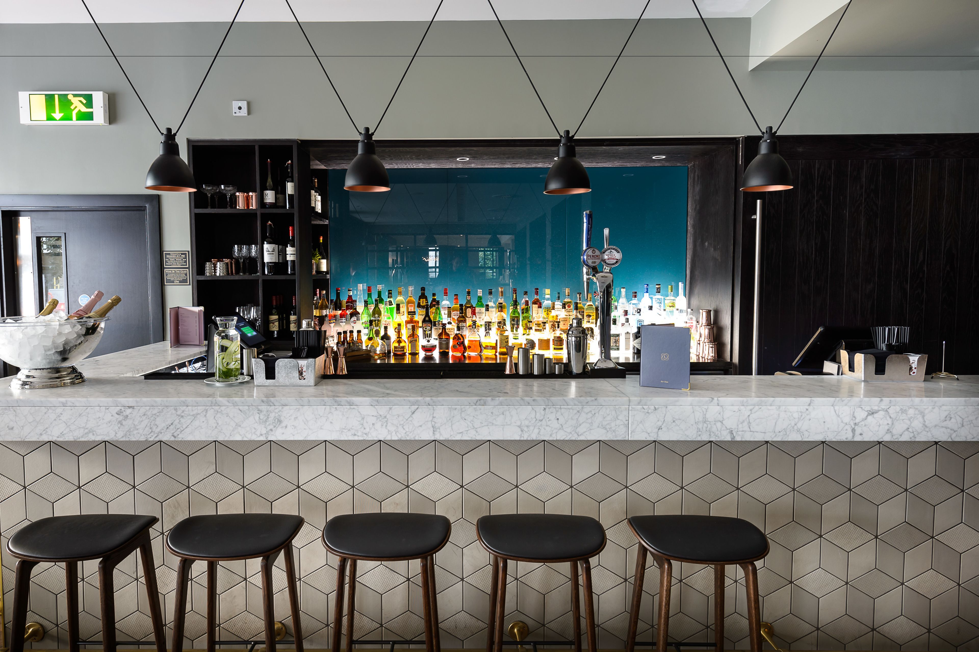 Una zona de bar con encimera de mármol, varias lámparas colgantes, una variedad de botellas y vasos, y una pared de azulejos con diseños geométricos.