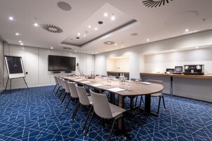 Sala de conferência com uma mesa comprida, cadeiras, quadro branco, TV de tela plana e um espaço para café.