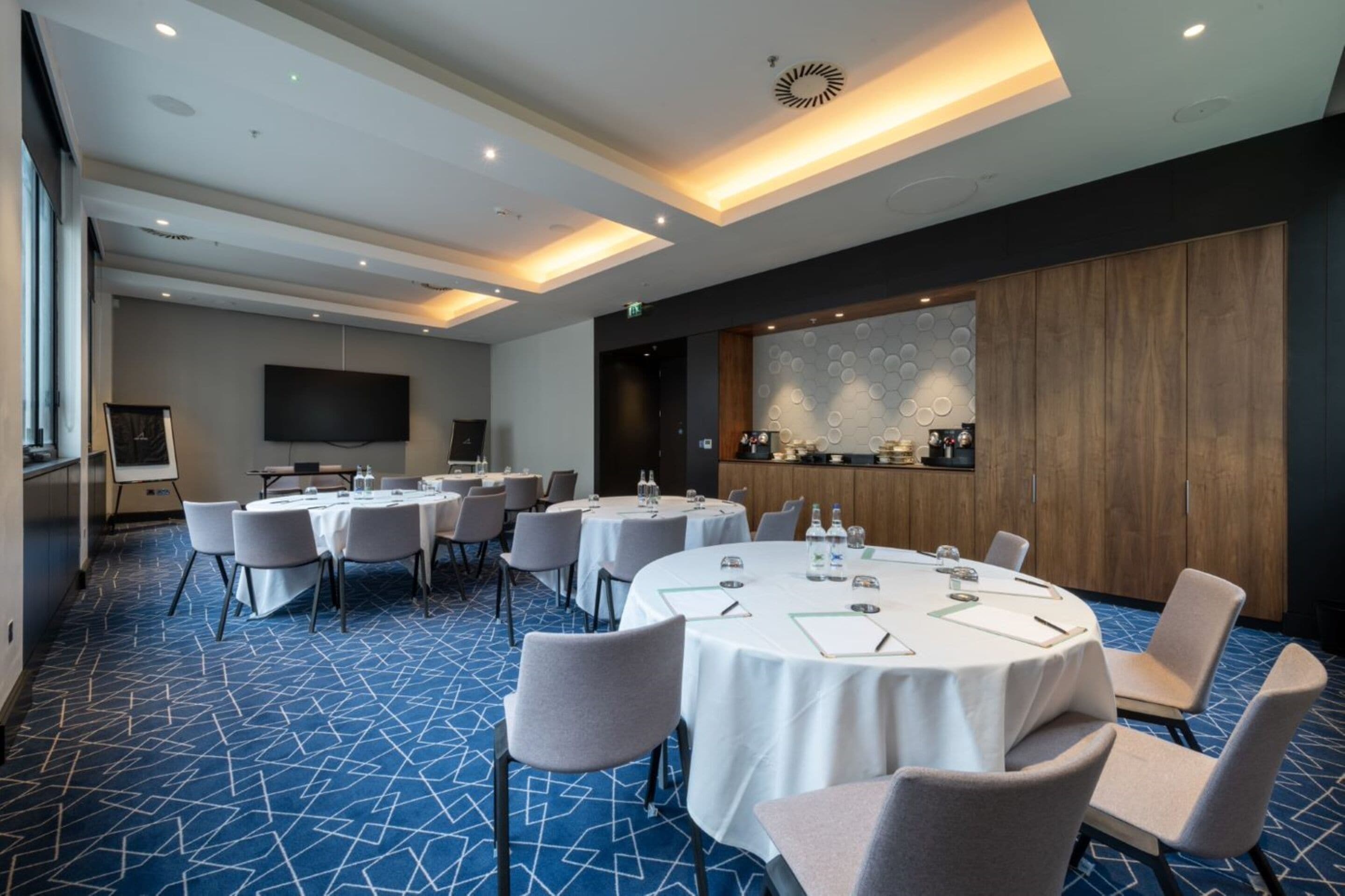 A conference room with multiple round tables, white tablecloths, and grey chairs. There is a large flat-screen TV mounted on the wall, a bar area with a wooden cabinet, and a patterned wall decoration.