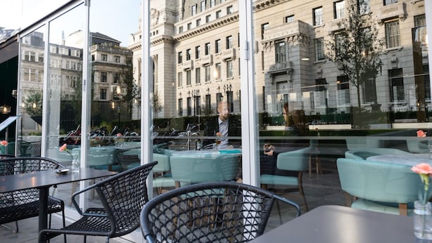An outdoor seating area with tables and chairs, large windows reflecting a historic building, and a clear sky. - Apex City of London Hotel (London)