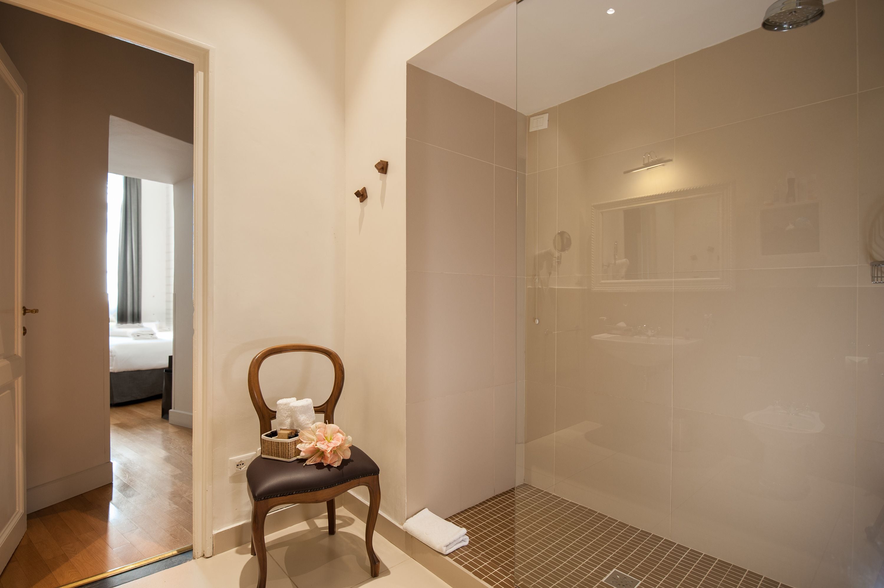 A modern bathroom with a glass-enclosed shower, a wooden chair with a tissue and a flower, and a visible bathroom counter with a mirror.