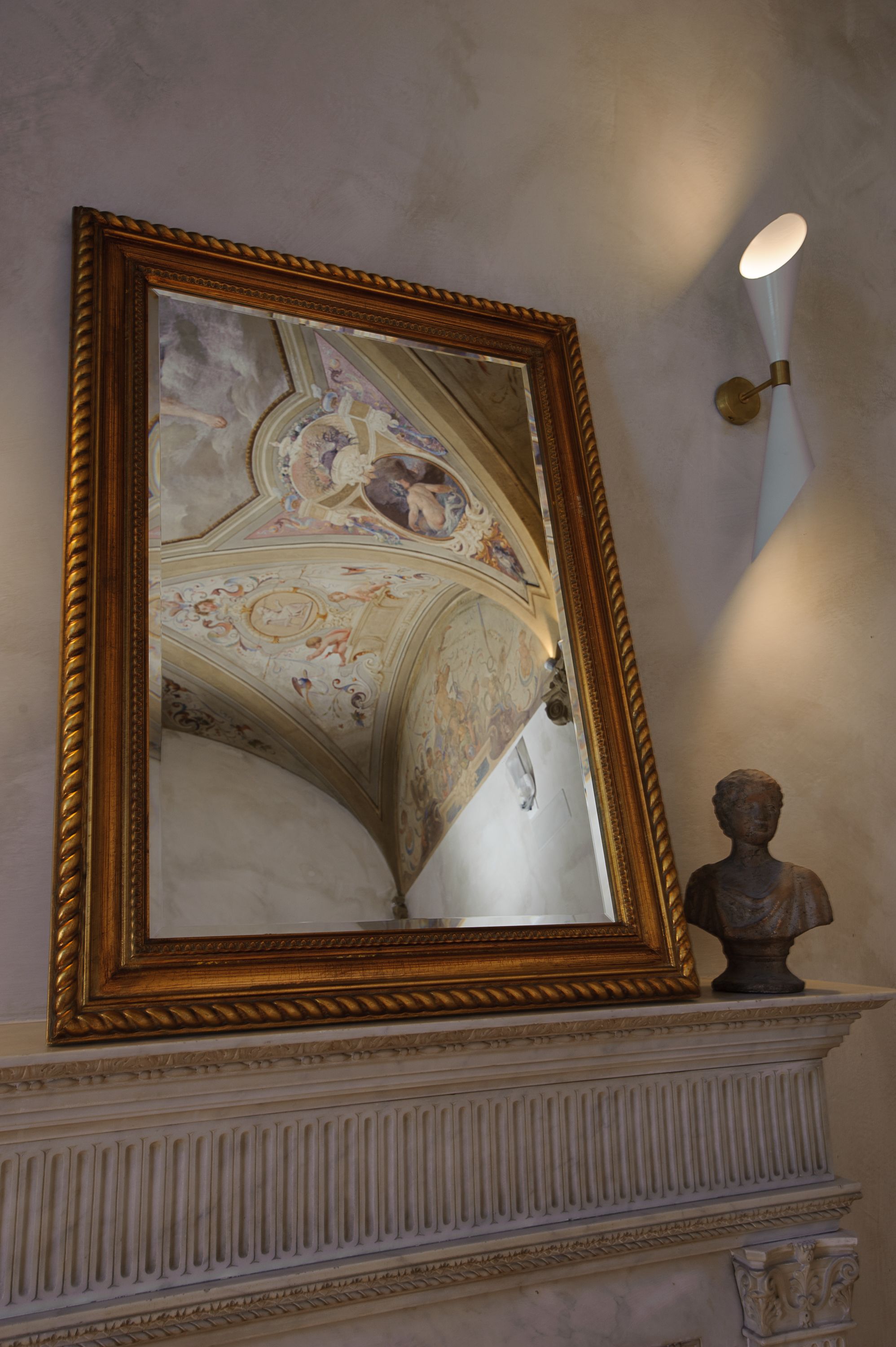 Un espejo en marco con el reflejo decorativo del techo, un aplique de pared y un busto en una repisa.