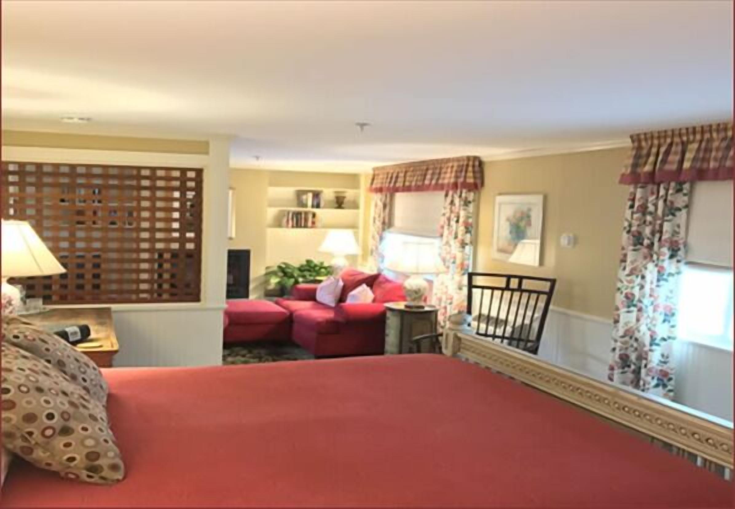 A hotel room with a large bed, a sofa, a small table with a lamp, a decorative screen, a window with curtains, and a bookshelf.