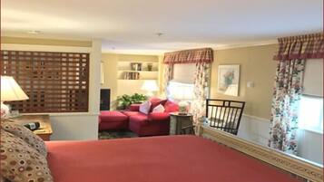 A hotel room with a large bed, a sofa, a small table with a lamp, a decorative screen, a window with curtains, and a bookshelf.