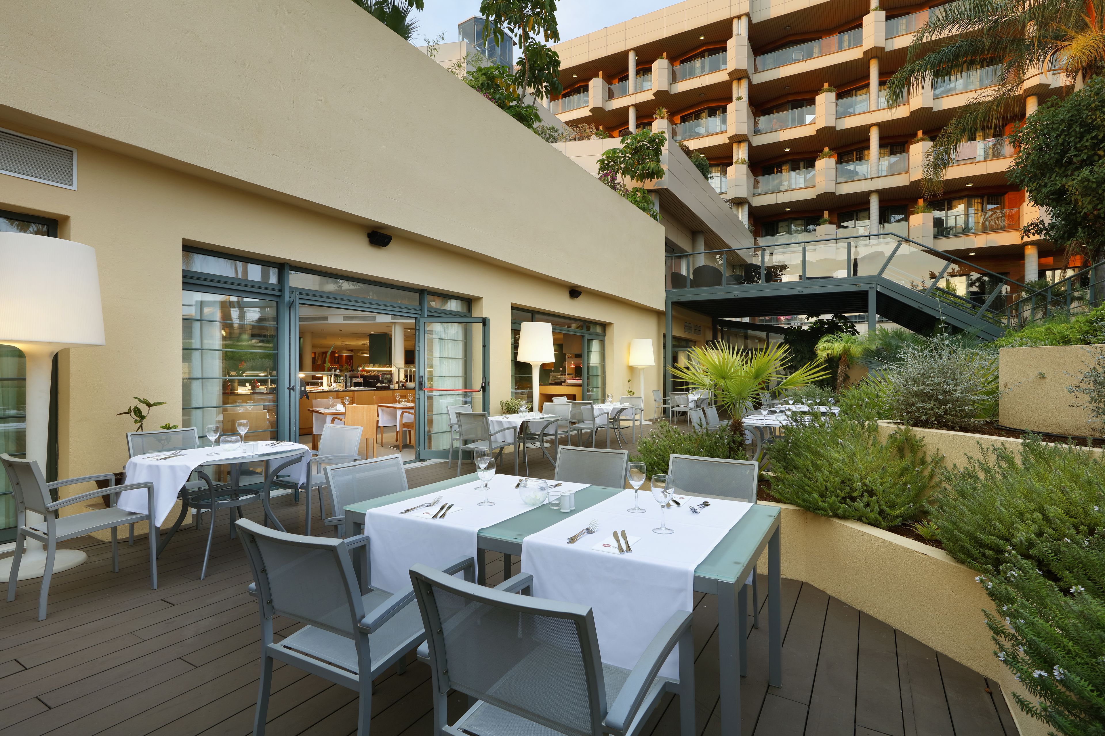 Un área de comedor al aire libre con mesas y sillas, un edificio con balcones y una escalera que lleva a un nivel superior.