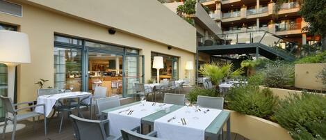 An outdoor dining area with tables and chairs, a building with balconies, and a staircase leading to an upper level.