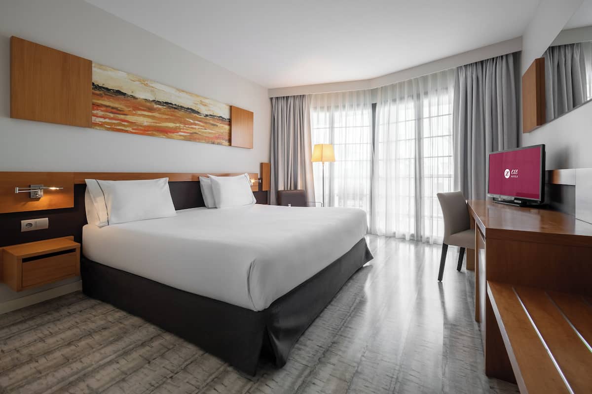 A modern hotel room with a large bed, a desk, a television, and a painting on the wall.
