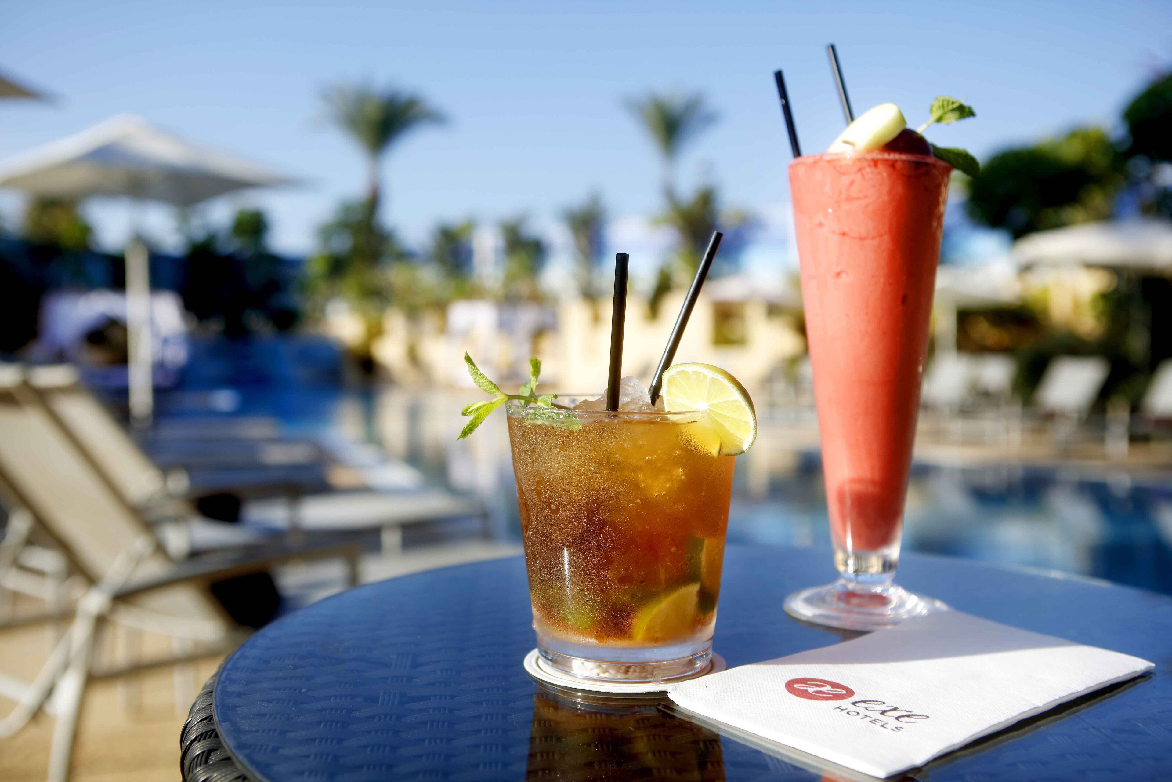 Two drinks on a table by a poolside, with a pool and lounge chairs in the background.