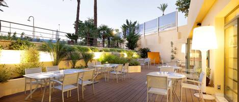 An outdoor patio with tables and chairs, surrounded by plants and palm trees.