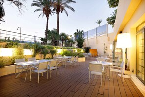 An outdoor patio with tables and chairs, surrounded by plants and palm trees.