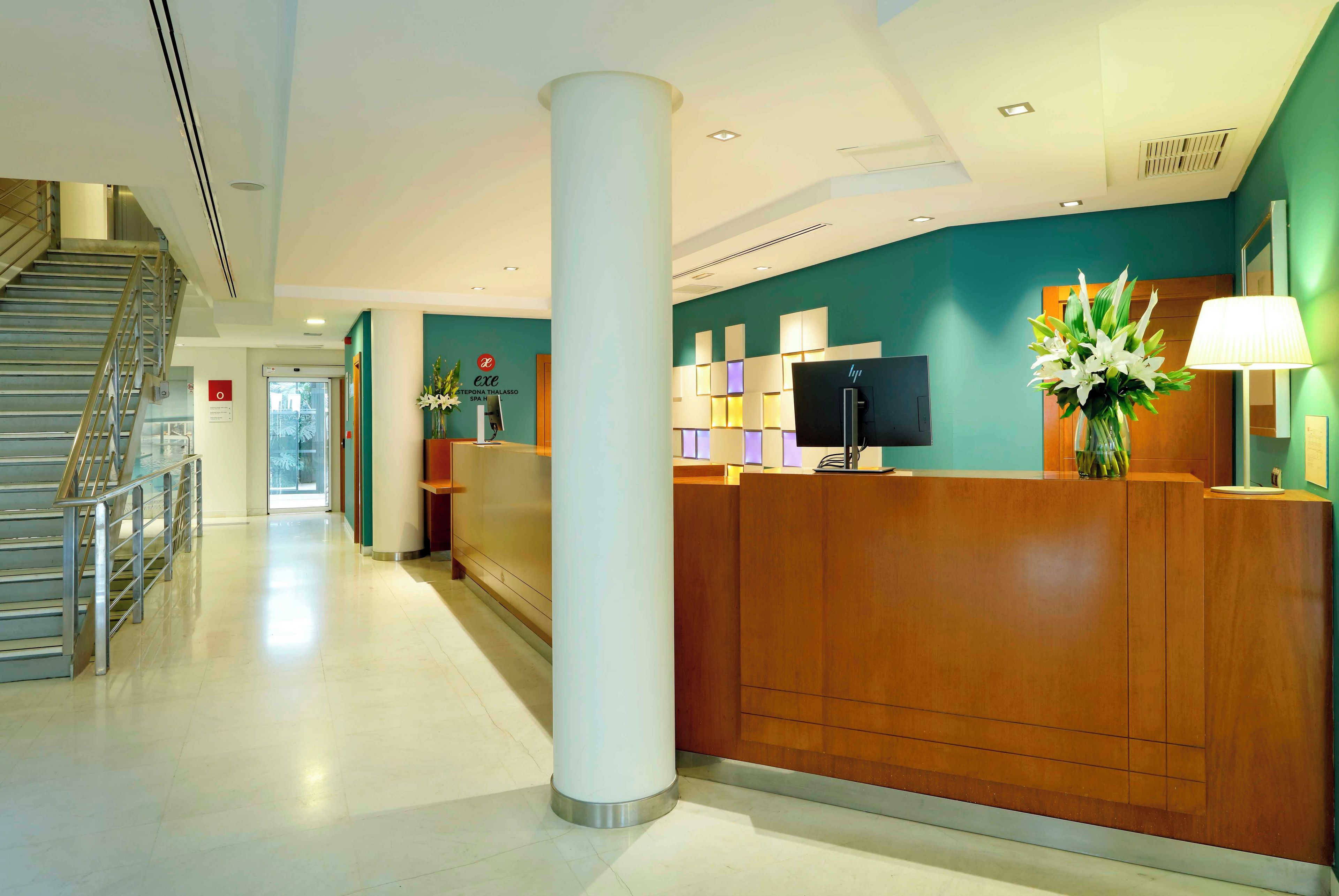 A hotel reception area with a wooden reception desk, a large pillar, a staircase, and a wall-mounted television.