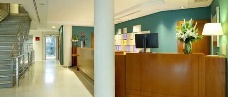A hotel reception area with a wooden reception desk, a large pillar, a staircase, and a wall-mounted television.