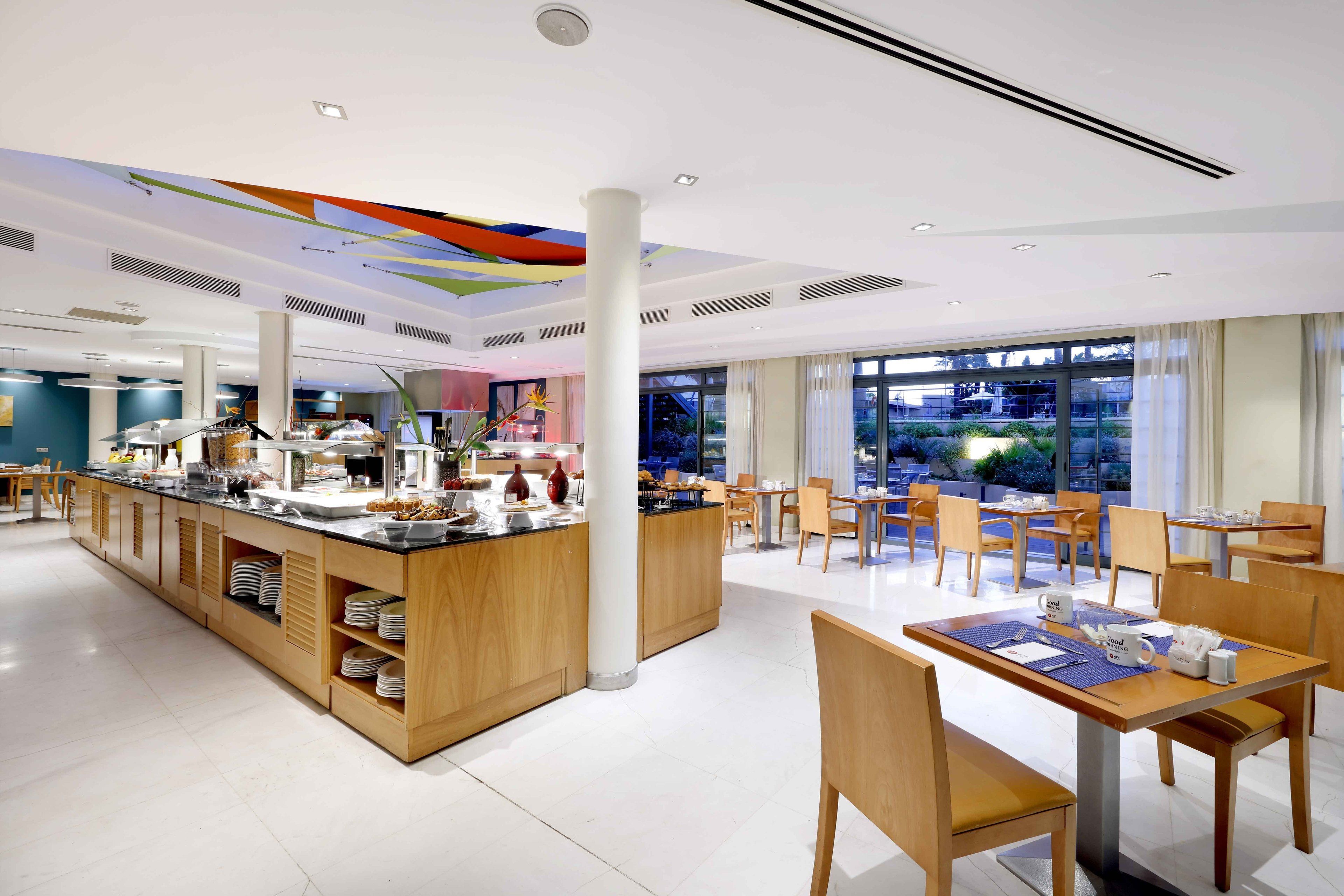 Un comedor de hotel moderno con un bufé central, mobiliario de madera y ventanas amplias que ofrecen vistas al exterior.