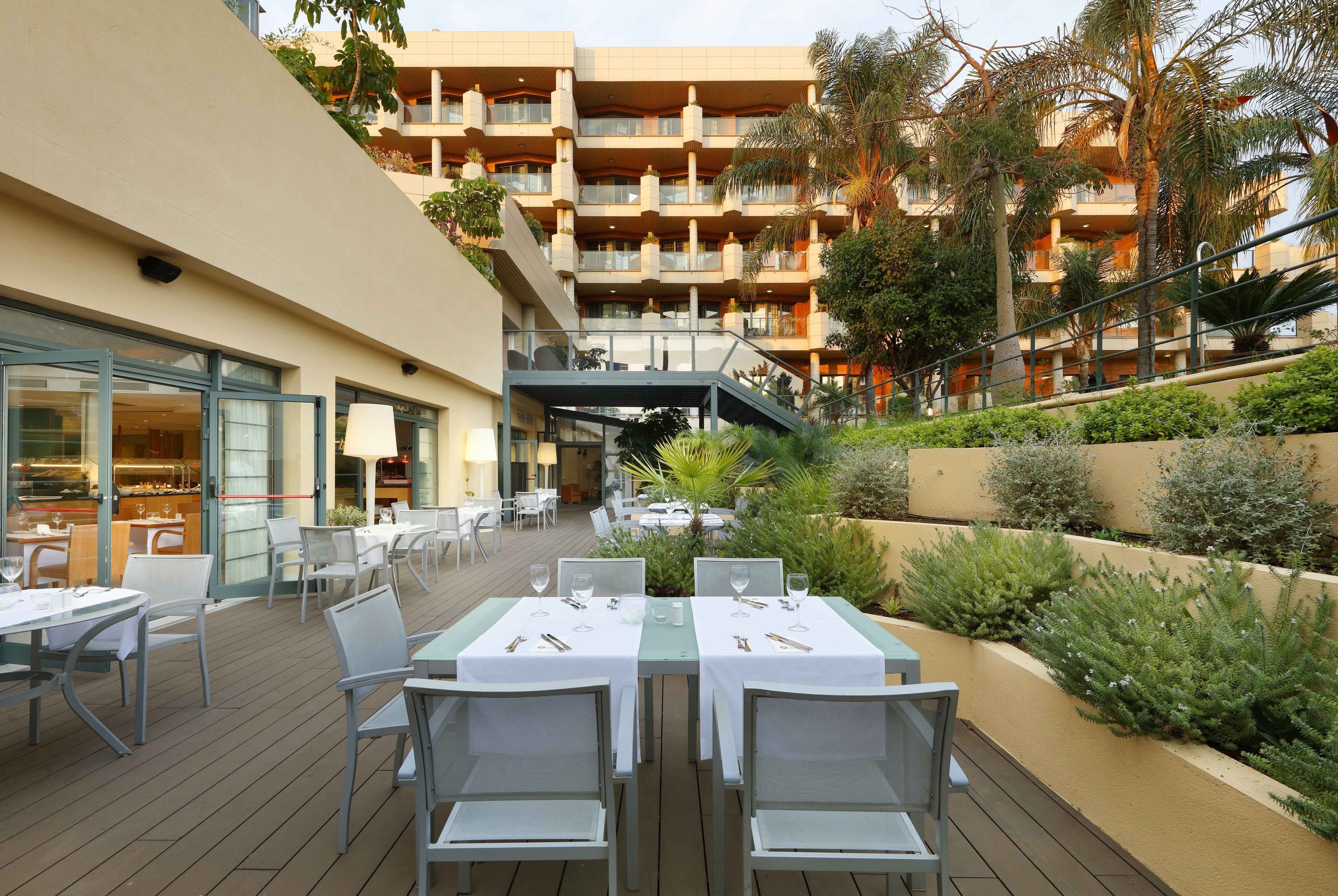 An outdoor dining area with tables and chairs, a restaurant on the left, and a multi-story building with balconies in the background.