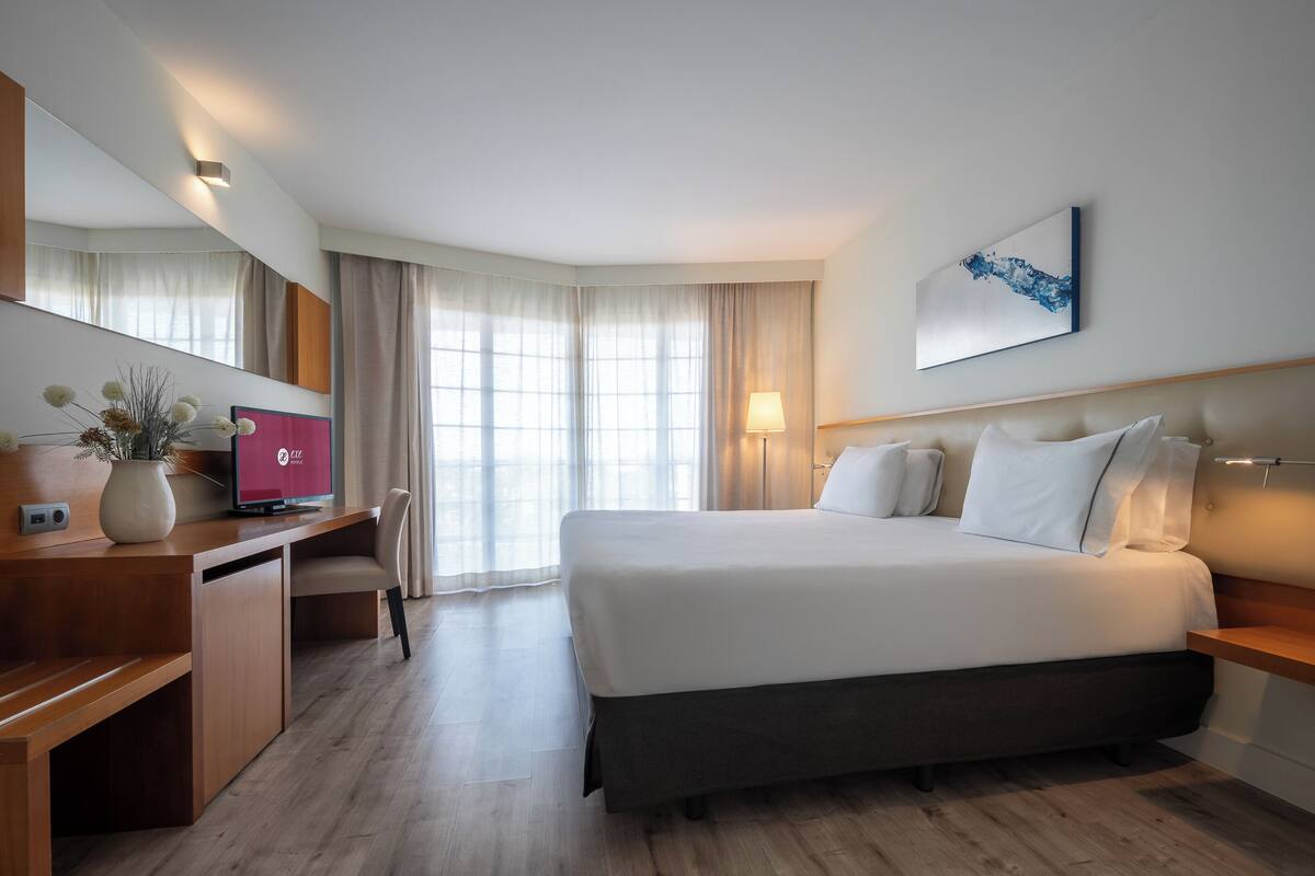 A modern hotel room with a large bed, a desk with a computer, a mirror, and a window with curtains.